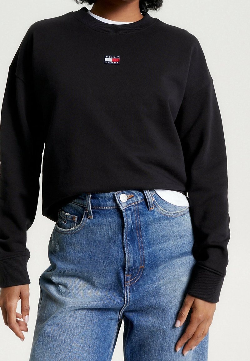 Sweatshirt nero accorciato con scollo rotondo e maniche lunghe, caratterizzato da un piccolo logo multicolore sul petto. Indossato con jeans blu a vita alta.