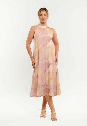 Kobieta w bezrękawowej pastelowej sukience midi tie-dye oraz beżowych sandałach na paski, stojąca z rękami na biodrach na białym tle.
