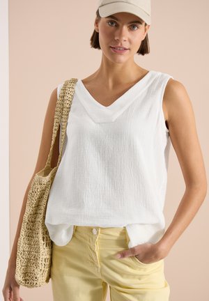 Femme portant un haut blanc sans manches, un pantalon jaune pâle, une casquette beige et portant un sac tissé beige sur l'épaule.
