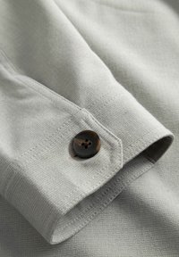 Tuvplāns gaiši pelēkas auduma piedurknes ar tumšu, četru caurumu pogu, kas aizpogāta apkašdaļā.