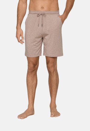 Man draagt beige knielange shorts met trekkoord in de taille, één hand in de zak, staat blootsvoets tegen een effen achtergrond.