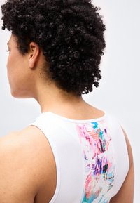 Γυναίκα με κοντά σγουρά μαλλιά που φοράει λευκό αμάνικο τοπ με πολύχρωμο αφηρημένο σχέδιο στην πλάτη πάνω σε απλό φόντο.