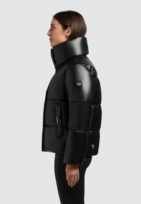 Juodas pūkinis švarkas su aukštu apykakle, siuvinėta konstrukcija ir blizgiu paviršiumi. Yra logotipo ženkliukas ant rankovės ir priekinė kišenė.