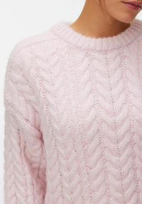 Femme portant un pull en tricot torsadé doux de couleur rose pâle, avec un col rond, montrant une texture de tricot tressé détaillée sur l'épaule et la poitrine.