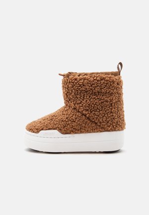 Bota de tobillo marrón borrosa con suela blanca gruesa y tira para tirar en la parte trasera sobre un fondo claro y liso.