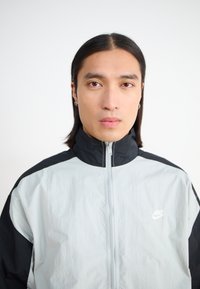 Nike vetrovka v svetlo sivi in črni barvi, ki ima visok ovratnik, sprednji zadrgo in logotip na levi prsih. Gladka in mehka tekstura tkanine.