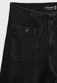 Jean noir taille haute avec fermeture à boutons à l'avant, rabat de poche latérale fixé par un bouton-pression noir, et étiquette de marque Stradivarius à l'intérieur de la ceinture.