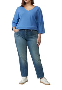 Blauwe gebreide trui met V-hals, gecombineerd met lichtgewassen jeans met rechte pijpen en witte sneakers met bruine accenten.