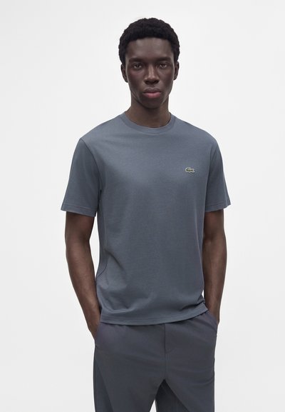 Jeune homme portant un t-shirt gris uni Lacoste avec un petit logo crocodile vert, debout les mains dans les poches sur un fond blanc.