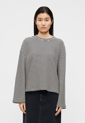 Pruhovaný svetr v černé a bílé barvě s dlouhými rukávy, kulatým výstřihem a volným střihem, ve spojení s tmavou sukní.
