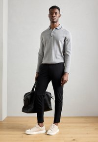 Grijze langekleurige polo, zwarte tapered broeken, beige sneakers en een zwarte sporttas, geplaatst op een houten vloer en een lichtgrijze achtergrond.