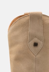 Botín de ante beige con forma redondeada, detalles de costura y un acento en forma de diamante con un logo de hardware cerca de la parte superior.