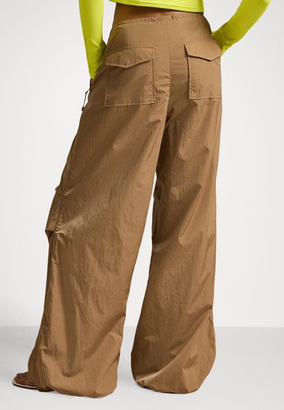 Persona valkā vaļīgas, platu kāju brūnas bikses ar lielām aizmugurējām kabatām un neona dzeltenu garām piedurknēm kreklu, stāvot ar rokām uz gurniem.