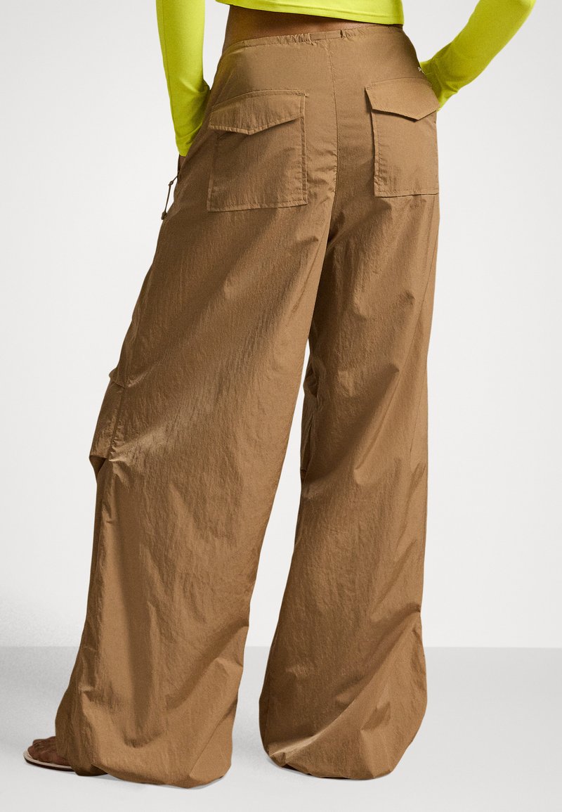 Person trägt lockere, weit geschnittene braune Hosen mit großen Gesäßtaschen und ein langärmeliges Neon-gelbes Oberteil, steht mit den Händen in den Hüften.