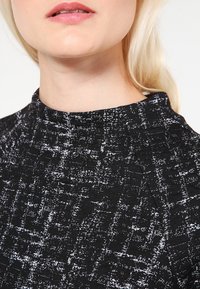 Svart texturerad topp med hög krage, prydd med ett vitt abstrakt mönster. Tyget verkar mjukt och något stretchigt.