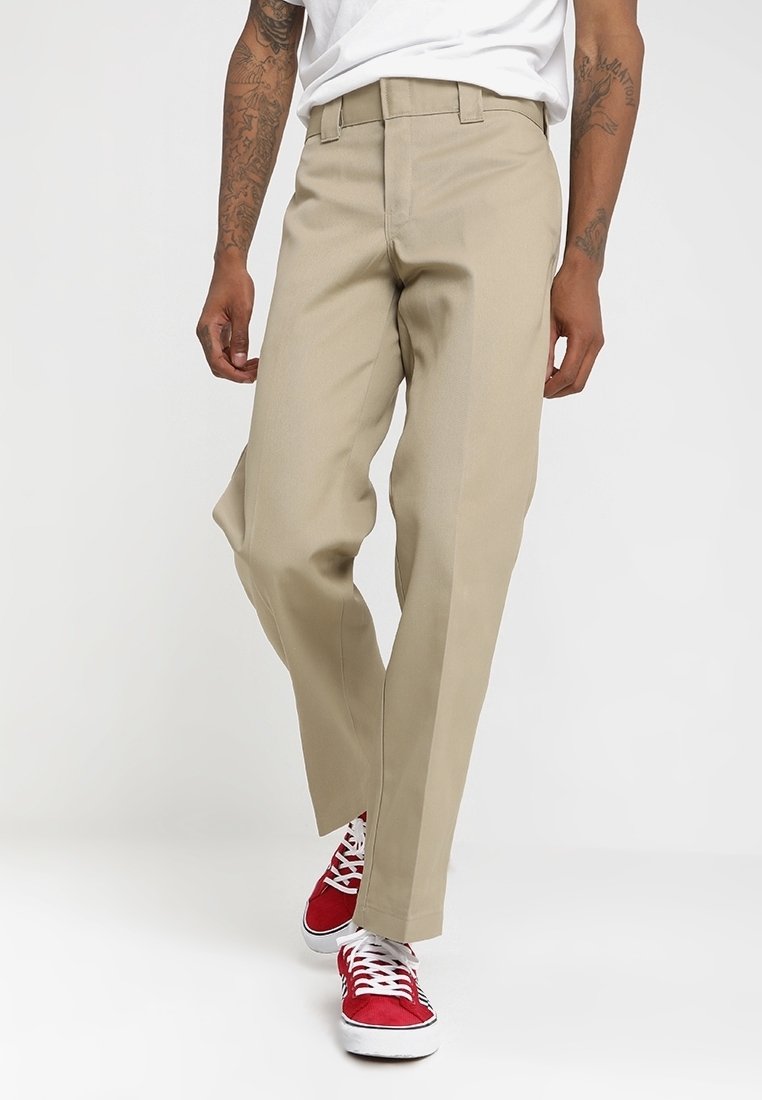 Osoba oblečená v béžových rovně střihových kalhotách a červených teniskách s bílými tkaničkami, stojící na jednobarevném bílém pozadí.