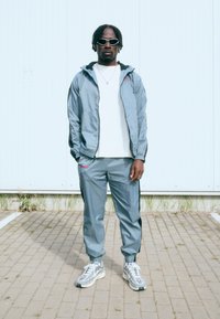 Lysblå træningsdragt med en lynlåsjakke og elastiske manchetter, parret med en hvid t-shirt og grå sneakers. Bemærkelsesværdige solbriller og kæde-accessory.