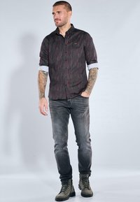 Schwarz-rot gestreiftes Hemd mit Knöpfen und hochgekrempelten Ärmeln, kombiniert mit dunkelgrauen Jeans und grünen Stiefeln, dargestellt auf einer stehenden Figur.