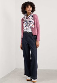 Jasnoróżowy dziergany kardigan, biała koszula, wzorzysty niebiesko-różowy szalik, ciemne dżinsy o szerokich nogawkach i białe tenisówki z jasnobrązową podeszwą.