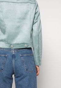 Personne portant une veste en daim vert clair et un jean bleu denim, vue de dos sur un fond uni.