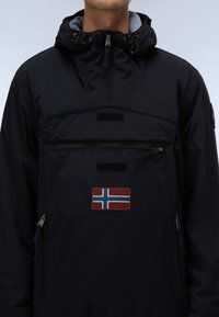 Mies, jolla on musta hupparillinen takki, jossa on etutasku vetoketjulla ja Norjan lipun merkki rinnassa.