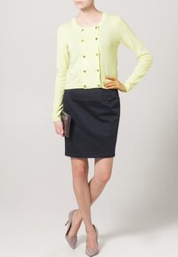 Helles gelbes Strickjacke mit goldenen Knöpfen, kombiniert mit einem schwarzen, figurbetonten Rock. Das Model ergänzt das Outfit mit einer dunklen Clutch und beigen Absatzschuhen.