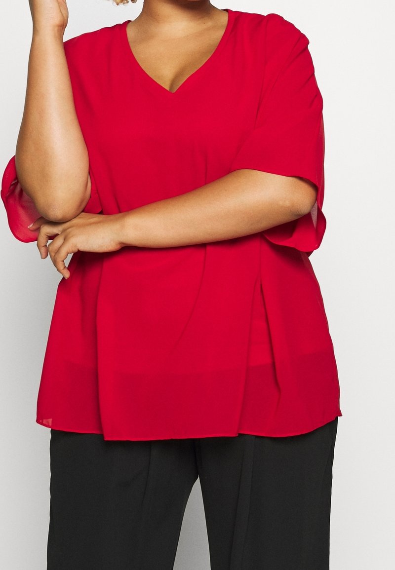 Blouse rouge à col en V, ample, en tissu léger, avec manches courtes et fentes latérales ; accompagnée d'un pantalon noir à jambes droites.