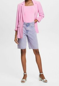 Blazer rosa sobre un top rosa sin mangas, combinado con bermudas de mezclilla azul claro y sandalias plateadas con tacón grueso. Tejido texturizado, corte entallado.