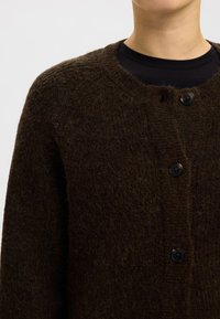 Sötétbarna kötött kardigán texturált, szőrös felülettel. Kerek nyakkivágással és fekete gombokkal a fronton, amelyekkel zárható.