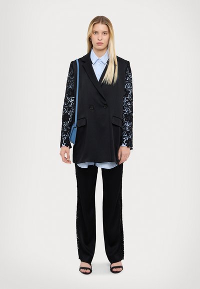Black sleeveless blazer with lace sleeves, layered over a light blue shirt, paired with black trousers featuring lace side panels and black sandals.