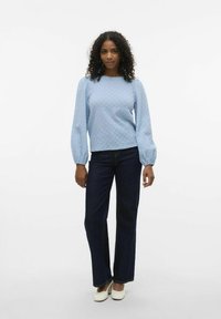 Blusa azzurro chiaro con texture e maniche a palloncino, abbinata a jeans svasati blu scuro. Il modello è in piedi su uno sfondo bianco uniforme.