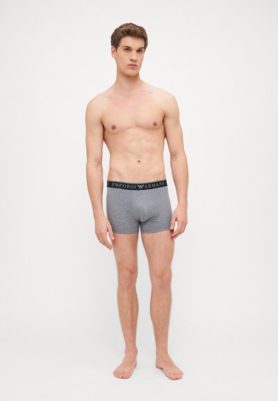 Graue Baumwollboxershorts mit einem schwarzen Bund, der das "Emporio Armani"-Logo zeigt, zu sehen an einem männlichen Model, das barfuß steht.