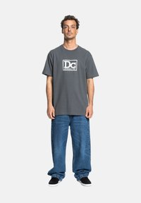 Graues Baumwoll-T-Shirt mit weißem Logo-Design auf der Brust, kombiniert mit lockeren blauen Jeans. Das Modell steht mit den Armen an den Seiten vor einem schlichten Hintergrund.