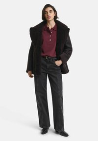 Woman wearing a burgundy polo shirt, black shearling jacket with wide collar, high-waisted black jeans, black belt, and black pointed shoes.