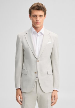 Homme aux cheveux bruns courts portant une veste et un pantalon de costume beige clair avec une chemise blanche, debout devant un fond clair uni.