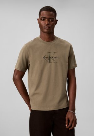 Νέος άνδρας που φοράει μπεζ μπλουζάκι Calvin Klein και σκούρο παντελόνι, στέκεται μπροστά σε απλό ανοιχτό γκρι φόντο.