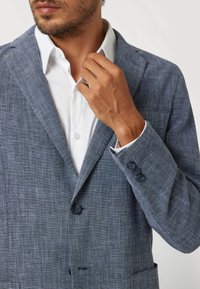 Tamsiai mėlynas tekstūruotas švarkas su išpjautu atvartėliu, vienu sagos užsegimu ir trimis sagomis ant rankogalių. Dėvimas virš baltos marškinėlių su apykakle.