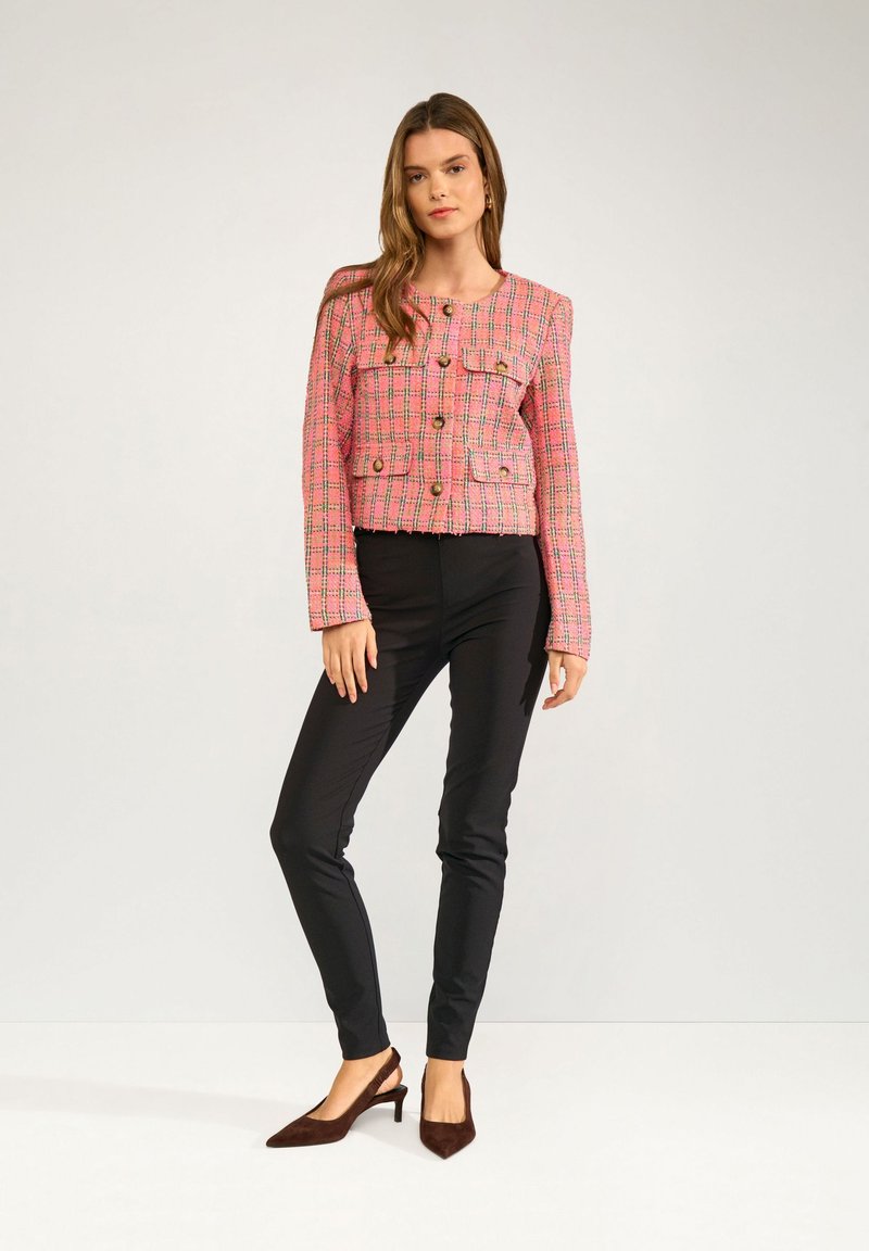 Frau steht, trägt eine rosa karierte Knopfleiste Jacke, schwarze enge Hose und braune spitze Slingback-Absätze vor einem schlichten hellen Hintergrund.