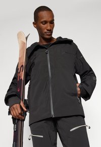 Zwarte waterdichte jas met een ritssluiting aan de voorkant, verstelbare capuchon, meerdere zakken en geprofileerde accenten. Het model houdt een ski vast en draagt een bijpassende broek.