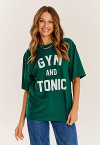 Zelené oversized tričko s nápisom "GYM AND TONIC" v bielej farbe. Krátke rukávy, pohodlný strih, vyrobené z mäkkej textílie, skombinované s modrými džínami.