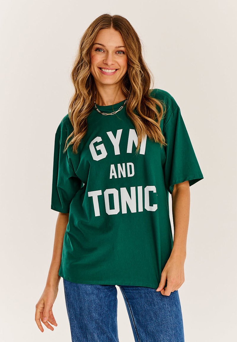 Zelené oversized tričko s nápisom "GYM AND TONIC" v bielej farbe. Krátke rukávy, pohodlný strih, vyrobené z mäkkej textílie, skombinované s modrými džínami.