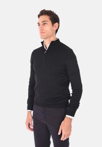 Maglione nero con zip e alto collo, orlo e polsini a coste, realizzato in tessuto liscio, indossato sopra una camicia bianca. Silhouette aderente.