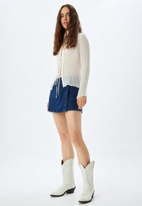 Blouse blanche transparent avec des plis verticaux et un détail à lacets, associée à une mini jupe en denim bleu et des bottes de cow-boy blanches.