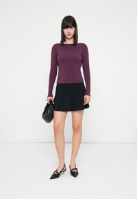 Maglioncino a maniche lunghe di colore viola abbinato a una gonna nera plissettata. La modella porta una piccola borsa nera e indossa delle ballerine nere a punta.