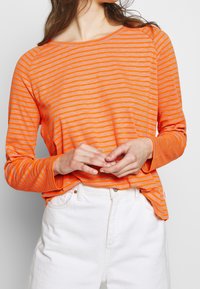 Långärmad topp i ljus orange med tunna horisontella ränder, med rund halsringning och avslappnad passform, kombinerad med vita byxor.