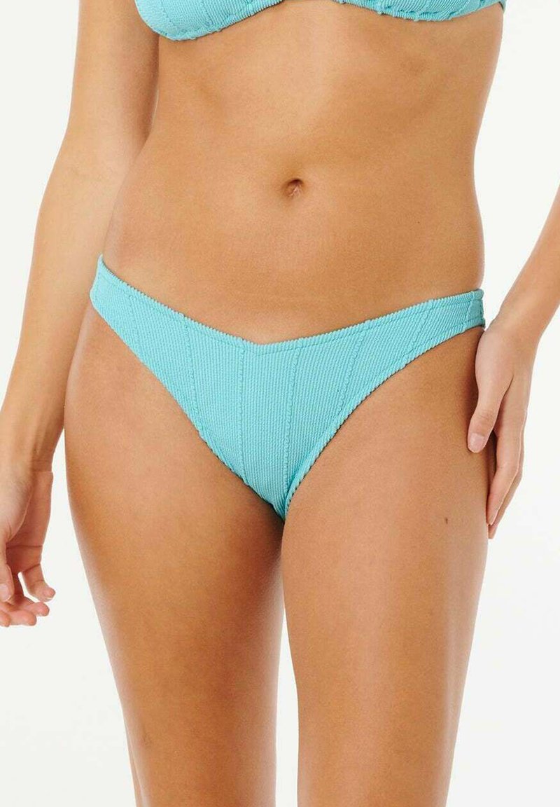 Kobieta w teksturowanych jasnoniebieskich majtkach bikini z przodem w kształcie litery V oraz pasującym staniku bikini, stojąca na jednolitym tle.