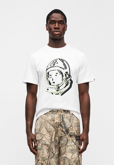 Λευκό βαμβακερό μπλουζάκι με γραφικό αστροναύτη σε μαύρο και πράσινο στο μπροστινό μέρος. Συνδυασμένο με μπεζ παραλλαγής παντελόνι cargo με υφασματικά μοτίβα.