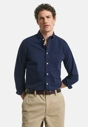 Marineblauw overhemd met knopen en een zak, met lange mouwen die iets opgerold zijn, in combinatie met khaki broek en een bruine riem.