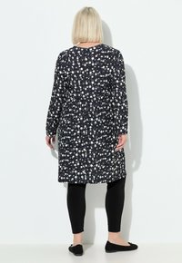 Kobieta z blond bobem, ubrana w długą tunikę w czarno-białe wzory, czarne legginsy i czarne baleriny, stojąca przy białej ścianie.