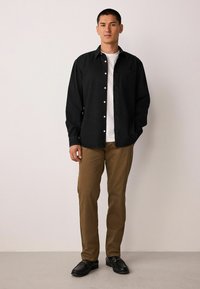 Mustast nööpidega särk valge t-särgi peal, beežid püksid ja mustad loafersid. Särgil on tasku ja lõdva istuvusega lõige.